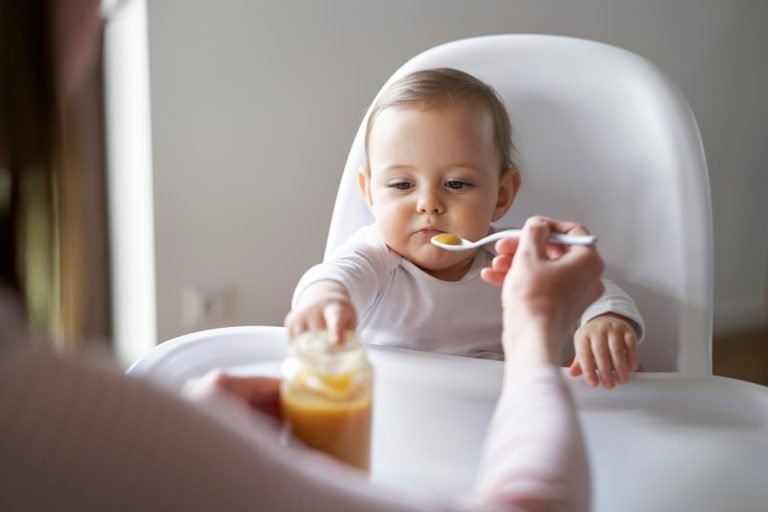Mom,Feeding,Baby,Girl,In,High,Chair,At,Home